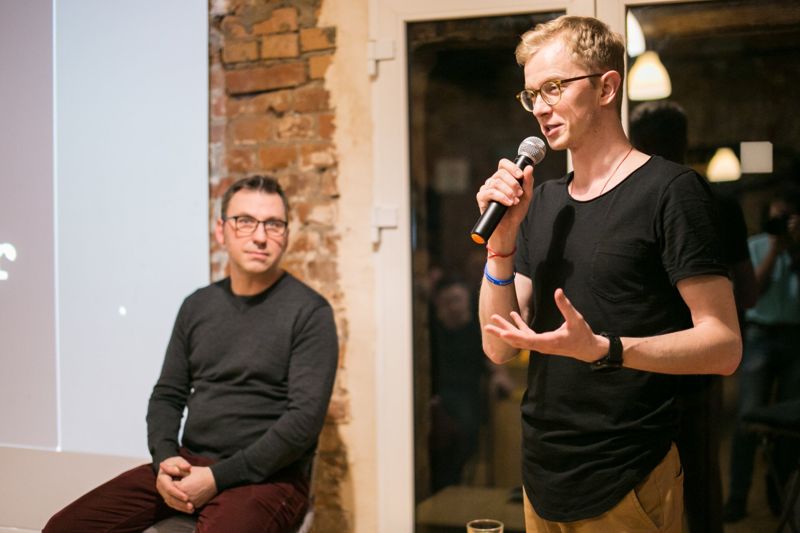 A man with a microphone speaking, another seated beside the screen