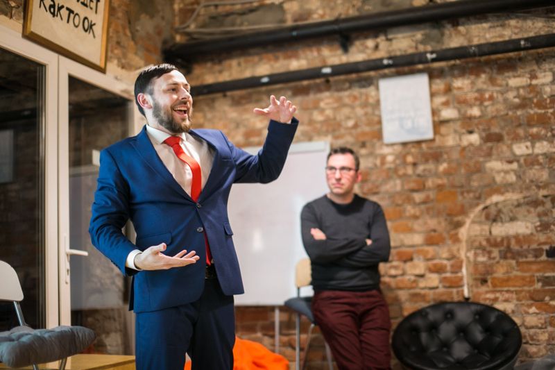 A founder in a blue suit and red tie, mid-pitch, arms wide, in a brick-walled room